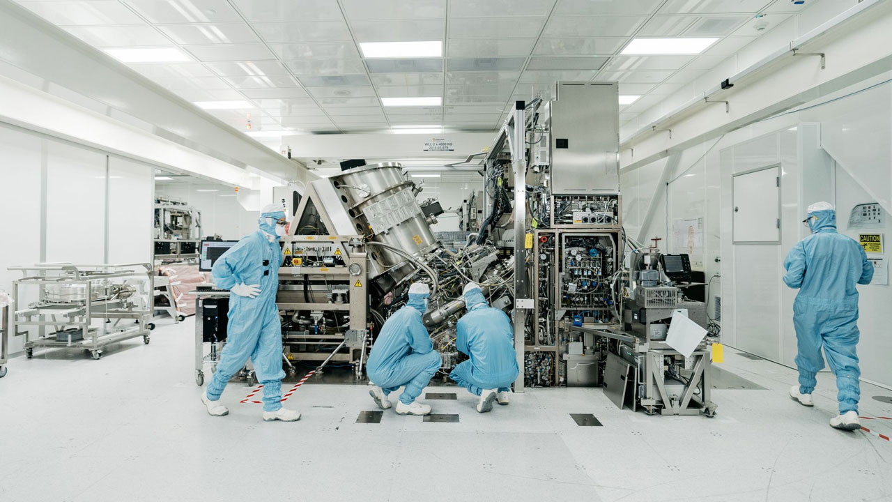 Inside a TSMC clean room, engineers in full white coveralls work beside large lithography equipment under bright filtered light.